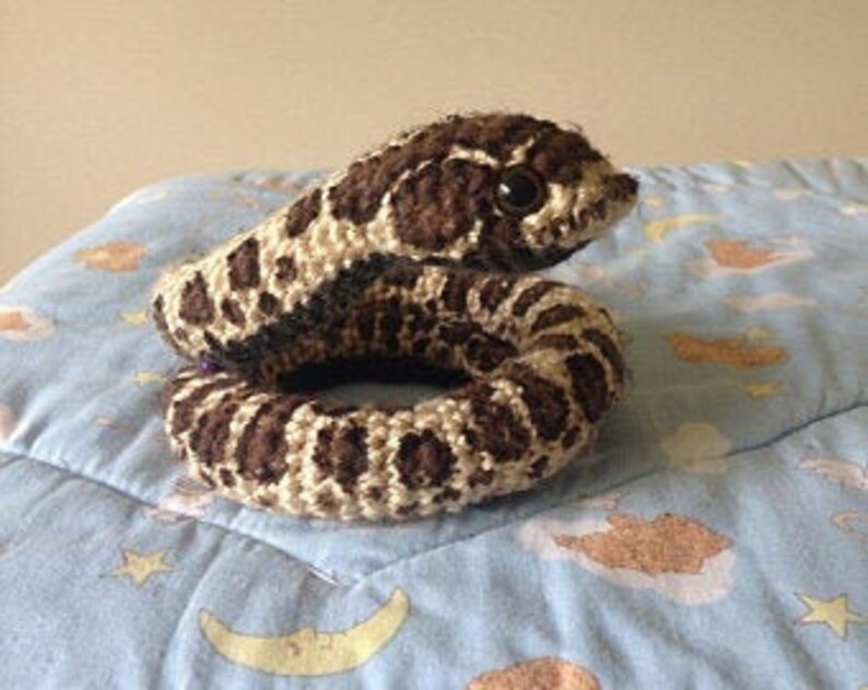 May include: A crocheted snake toy in shades of brown and cream. The snake is coiled, displaying a detailed spot pattern and a small, dark eye. It rests on a blue patterned surface.