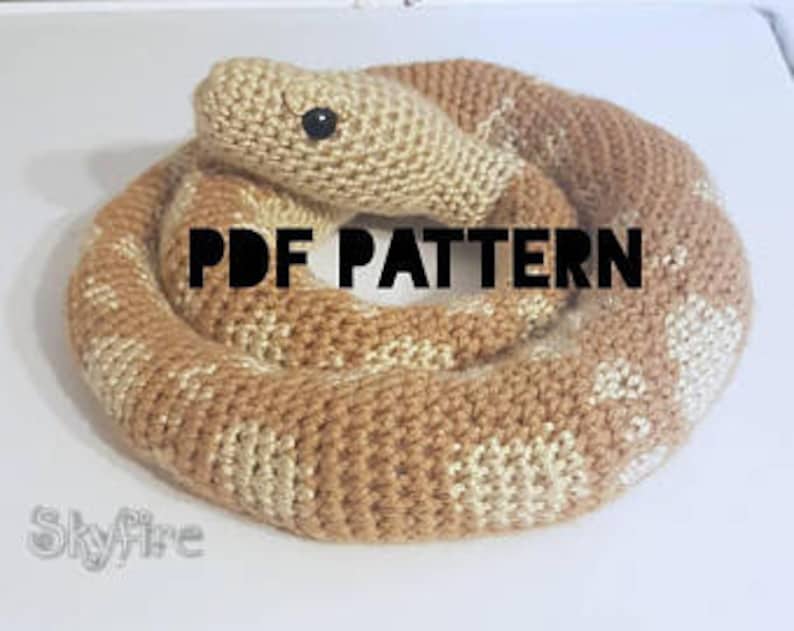 May include: A knitted snake toy in shades of tan and cream, coiled on a white surface. The snake has a detailed pattern and a black eye. The text "PDF PATTERN" is displayed in black. The word "Skyfire" is also visible.