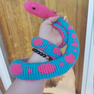 以下が含まれることがあります： 明るい色の手作りの編み物ヘビのおもちゃ。ヘビの頭はピンク色で、体はターコイズブルーでピンク色のハート型の模様があります。おもちゃは柔らかく、糸で作られているようです。