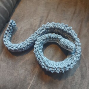 Op de afbeelding: Een blauwgrijs gehaakt slangenknuffel, opgerold op een bruin oppervlak. De slang heeft een getextureerd lichaam en een kleine kop met zwarte ogen. Dit handgemaakte item is een grillig knutselwerk.