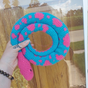 May include: A turquoise crocheted snake with pink heart-shaped spots. The head and tail are solid pink. The snake is coiled. The toy is handmade and appears soft. The snake is a decorative item.