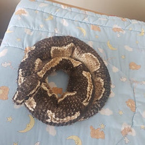 以下が含まれることがあります： 茶色、ベージュ、クリーム色の鉤針編みのヘビのおもちゃが、月と星の模様が入った水色の毛布の上に丸まっています。ヘビの質感がよくわかり、ステッチも細かく施されています。