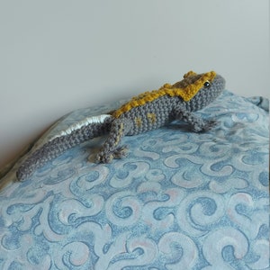 May include: A handmade crocheted lizard toy in grey and yellow, with a white tail tip. The reptile is lying on a blue patterned surface. The lizard's body is detailed with crocheted scales and small legs.