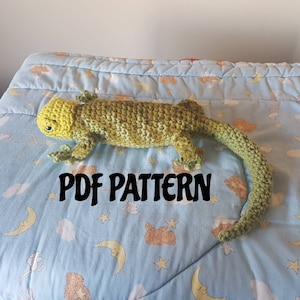 Puede incluir: Un lagarto de ganchillo hecho a mano en tonos verdes y amarillos, sobre una manta azul claro estampada. El lagarto tiene una cola larga y patas detalladas. El texto "PDF PATTERN" es visible.