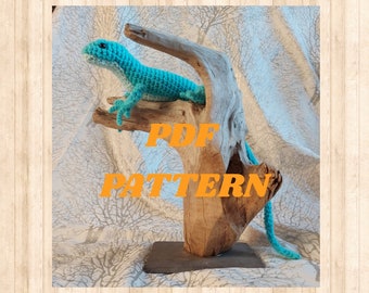 May include: A teal blue crocheted lizard sitting on a piece of driftwood. The driftwood is on a black slate base. The text "PDF PATTERN" is written in orange letters on the driftwood.