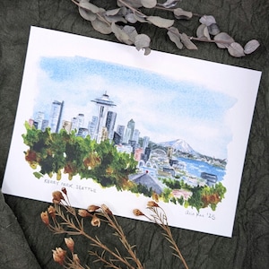 May include: Watercolor painting of Seattle's skyline, including the Space Needle, buildings, and a mountain. The artwork is signed "Alia Rae '25" and titled "Kerry Park, Seattle". The painting features a blue sky and green trees.