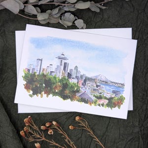 May include: Watercolor art print of the Seattle skyline, including the Space Needle, buildings, and a mountain range. The print is on white cardstock, with a blue sky and green trees. The art print is on top of a blank white card.