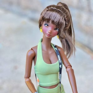 May include: A brown-skinned doll with long brown hair in a ponytail, wearing a green athletic outfit and a yellow backpack, holds the handlebars of a scooter.