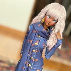 May include: A doll wearing a navy blue coat dress with a floral pattern in shades of orange and red. The dress features large gold buttons, pockets, and a matching scarf. The doll has long, light pink hair and is wearing black knee-high socks.