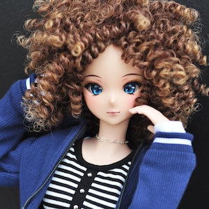 May include: A doll with large blue eyes and curly brown hair. The doll wears a blue zip-up jacket over a black and white striped top. A silver necklace and a skirt with a dog pattern complete the outfit.