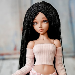 May include: A doll with long black braids, wearing a light pink off-the-shoulder top and heart-patterned leggings. The doll has detailed facial features and is posed in a studio setting.