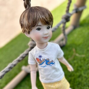 May include: A doll with brown hair wearing a white t-shirt with a blue and orange graphic that says "Ride the Waves". The doll is wearing yellow shorts and is standing on a green grassy surface.