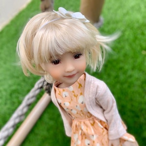 May include: A blonde doll wearing a peach dress with a floral pattern and a light pink cardigan. The doll is wearing white shoes and has a white bow in her hair.
