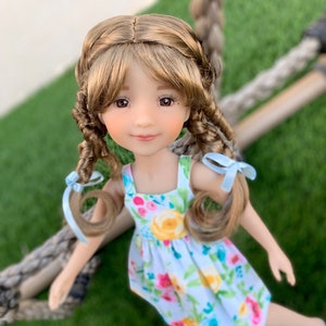 May include: A doll with long brown hair styled in braids with blue ribbons. She is wearing a white dress with a floral pattern and is standing on a green grassy surface.