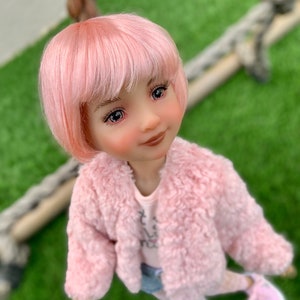 Puede incluir: Una muñeca rosa con cabello rosa, que lleva una chaqueta de peluche rosa, una camisa blanca con un diseño rosa, pantalones cortos de mezclilla azul y zapatos rosas.
