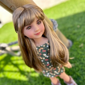 Puede incluir: Una muñeca con el pelo largo y castaño que lleva un vestido verde con flores blancas y naranjas. La muñeca lleva botas marrones y está de pie sobre hierba verde.