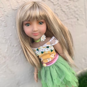 May include: A doll with long blonde hair wearing a yellow and green dress with floral accents and a green tulle skirt. She is wearing a green and white flower choker and has green eyes.