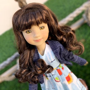 May include: A doll with long brown curly hair, wearing a blue cardigan and a light blue dress with a colorful pattern. The doll has brown eyes and a light complexion.