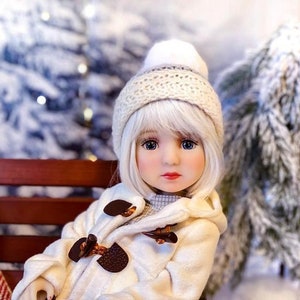 May include: A white doll with blonde hair wearing a white knitted hat and a white coat with brown buttons. The doll is sitting on a wooden bench in front of a snowy background.