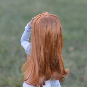 May include: A young girl with long, straight, red hair is wearing a light blue shirt with a white pattern. She is standing in a grassy field.
