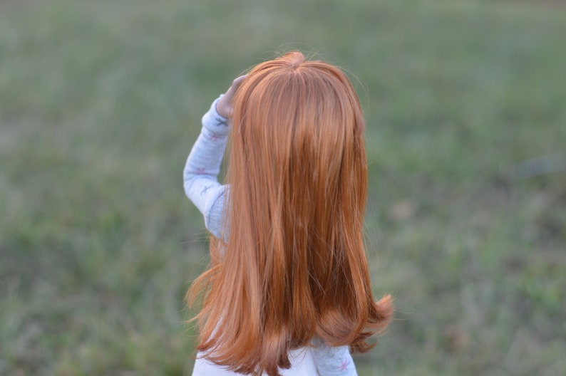 May include: A young girl with long, straight, red hair is wearing a light blue shirt with a white pattern. She is standing in a grassy field.