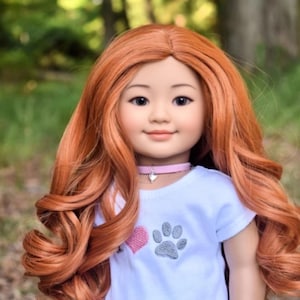 May include: A doll with long, curly, auburn hair wearing a white t-shirt with a pink heart and gray paw print design. The doll is also wearing a gray and pink skirt with a paw print and heart pattern. The doll is wearing a pink choker necklace and a pink bracelet.