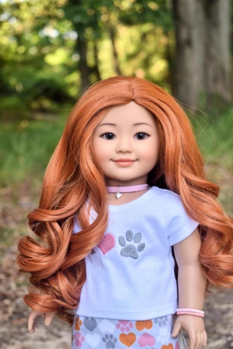 May include: A doll with long, curly, auburn hair wearing a white t-shirt with a pink heart and gray paw print design. The doll is also wearing a gray and pink skirt with a paw print and heart pattern. The doll is wearing a pink choker necklace and a pink bracelet.