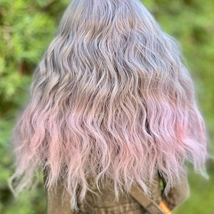 May include: A doll with long, wavy hair. The hair is a blend of gray and silver at the roots, transitioning to a soft pink at the ends. The doll is wearing a brown jacket with a strap across the chest.