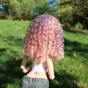 May include: A doll with long, curly, pink hair. The doll is wearing a white top and blue jeans.