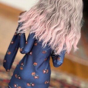 May include: A navy blue dress with a floral pattern in shades of orange and red. The dress has long sleeves and a rounded neckline. The model is wearing a wig with wavy gray hair that fades to pink at the ends. The model is also wearing black boots.