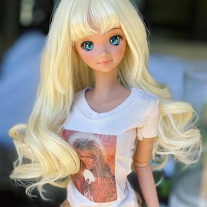 May include: A blonde doll with blue eyes wearing a white t-shirt with a printed image of a man with long hair. The doll is wearing brown shorts and has a long, wavy hairstyle.