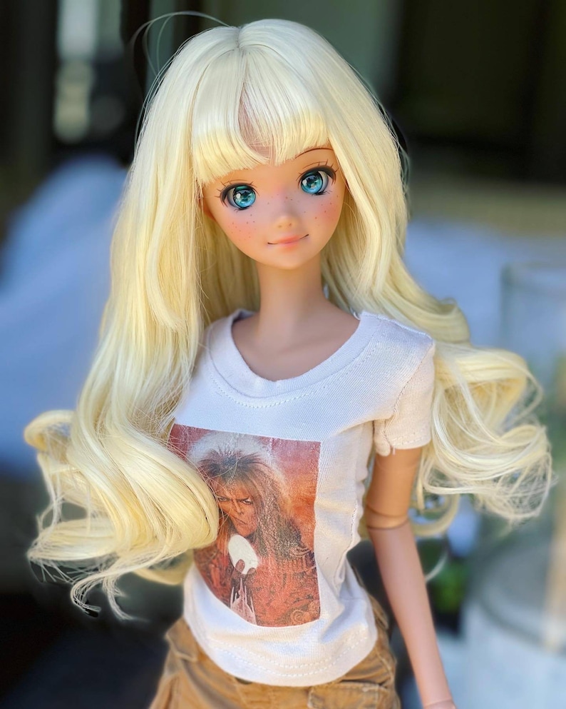 May include: A blonde doll with blue eyes wearing a white t-shirt with a printed image of a man with long hair. The doll is wearing brown shorts and has a long, wavy hairstyle.