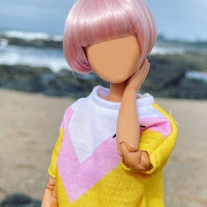 Peut inclure: Une poup&eacute;e aux cheveux roses portant un pull jaune, rose et blanc. La poup&eacute;e est debout sur une plage avec des rochers et de l'eau en arri&egrave;re-plan.