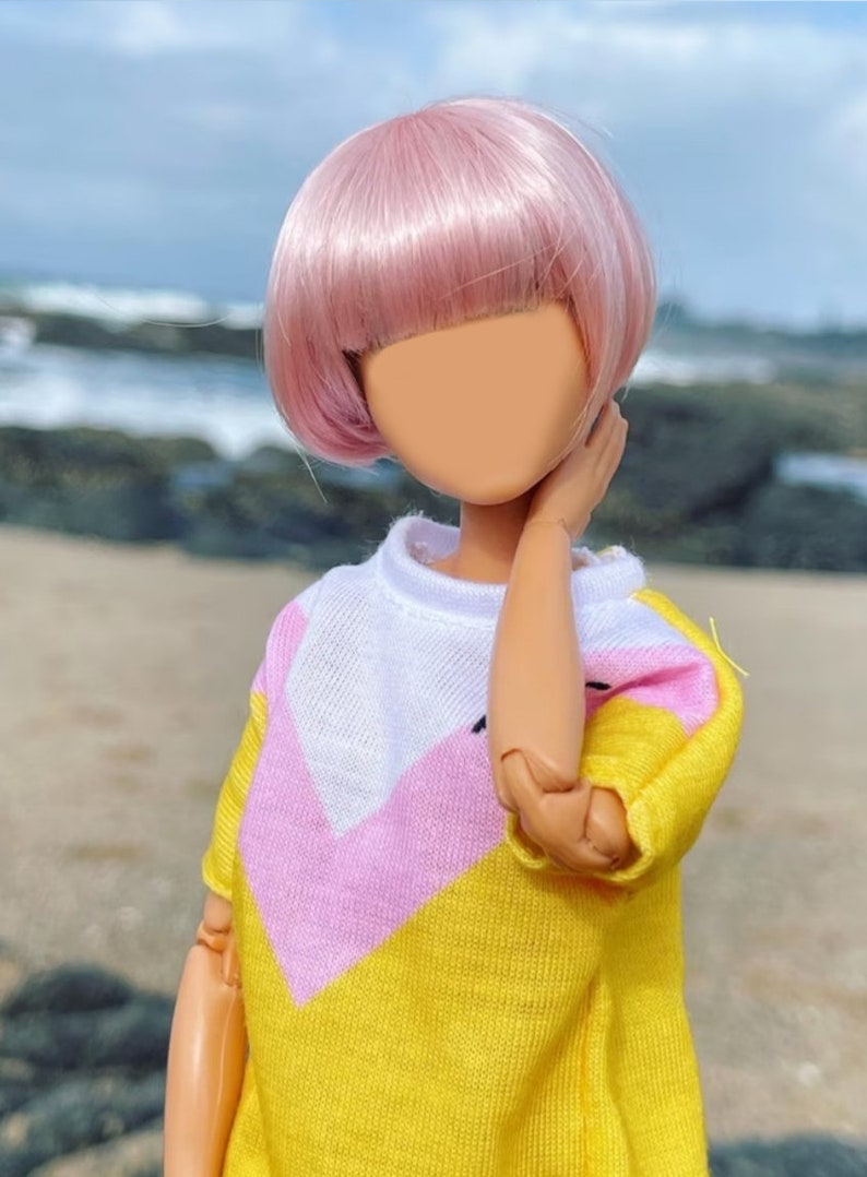 Peut inclure: Une poup&eacute;e aux cheveux roses portant un pull jaune, rose et blanc. La poup&eacute;e est debout sur une plage avec des rochers et de l'eau en arri&egrave;re-plan.