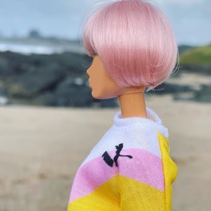 Peut inclure: Une poup&eacute;e aux cheveux roses portant une chemise ray&eacute;e jaune, rose et blanche avec un logo noir. La poup&eacute;e est debout sur une plage de sable.