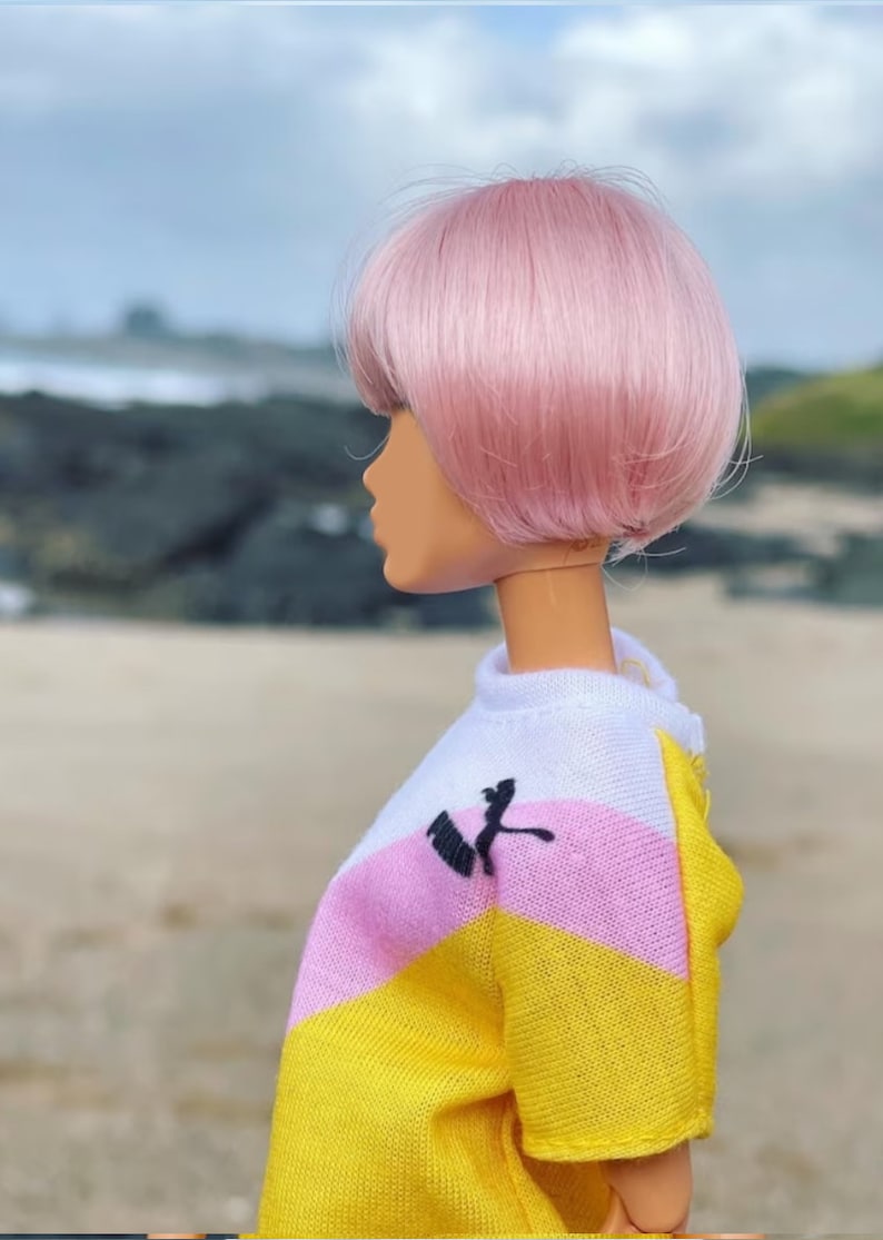 Peut inclure: Une poup&eacute;e aux cheveux roses portant une chemise ray&eacute;e jaune, rose et blanche avec un logo noir. La poup&eacute;e est debout sur une plage de sable.