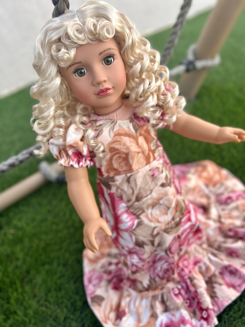 May include: A doll with blonde curly hair wearing a pink and brown floral dress.
