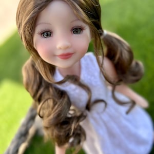 Könnte beinhalten: Eine Porzellanpuppe mit langen braunen Haaren, die zu Zöpfen geflochten sind, trägt ein weißes Kleid. Die Puppe hat realistische Gesichtszüge, darunter grüne Augen und ein sanftes Lächeln.