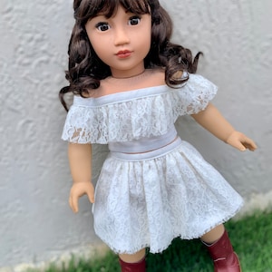 May include: A doll wearing a white lace off-the-shoulder top and skirt with brown boots. The doll has long brown hair with bangs and brown eyes.
