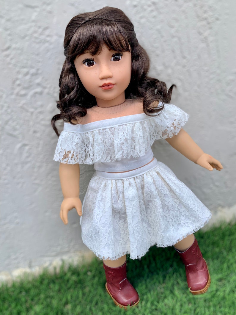 May include: A doll wearing a white lace off-the-shoulder top and skirt with brown boots. The doll has long brown hair with bangs and brown eyes.