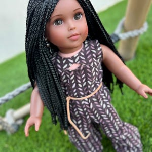 May include: A black doll with long black braids wearing a purple and white patterned jumpsuit with a brown belt.