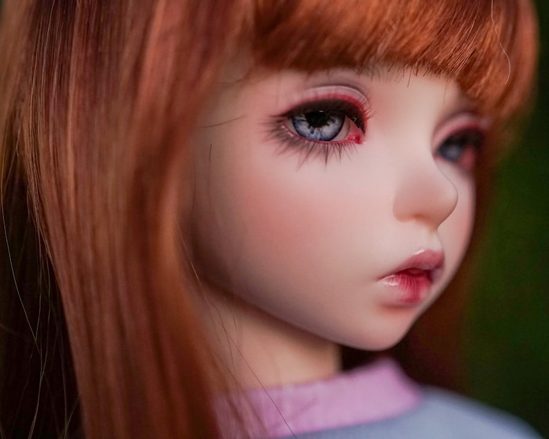 May include: A close-up of a doll's face with red eyeshadow, long eyelashes, and blue eyes. The doll has long, straight, red hair.