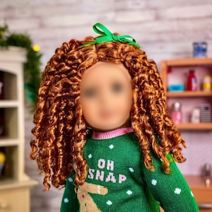 Peut inclure: Une poupée avec de longs cheveux roux bouclés, coiffés d'un nœud vert. La poupée porte un pull vert avec le texte "OH SNAP" et un motif de renne. L'arrière-plan comprend une cuisine.