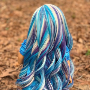May include: A doll with long, wavy hair in shades of blue, white, and purple. The hair is styled in a layered look, with the ends of the hair curling slightly.