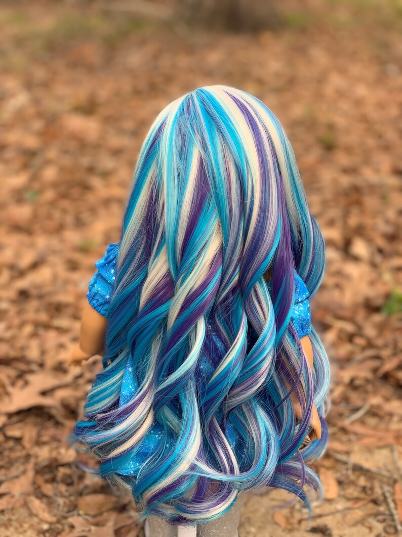 May include: A doll with long, wavy hair in shades of blue, white, and purple. The hair is styled in a layered look, with the ends of the hair curling slightly.