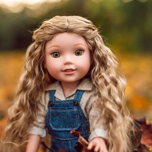 May include: A doll with long, curly blonde hair wearing a denim overall dress and a white shirt with small polka dots. The doll is sitting in a pile of autumn leaves.