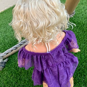 May include: A purple lace dress with a ruffled neckline and a floral pattern. The dress is worn by a doll with blonde curly hair.