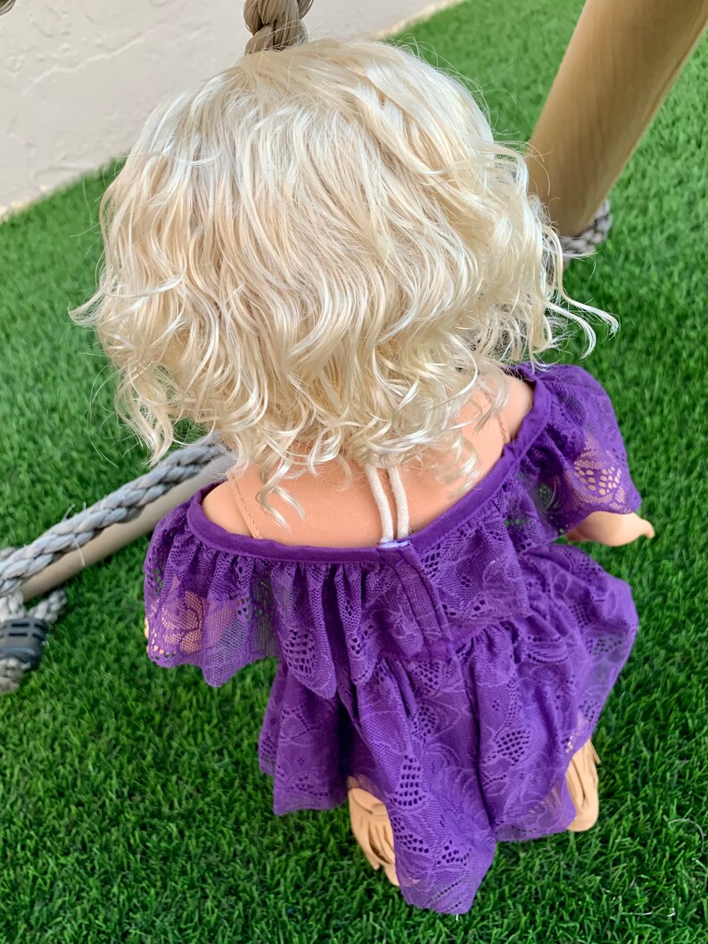 May include: A purple lace dress with a ruffled neckline and a floral pattern. The dress is worn by a doll with blonde curly hair.