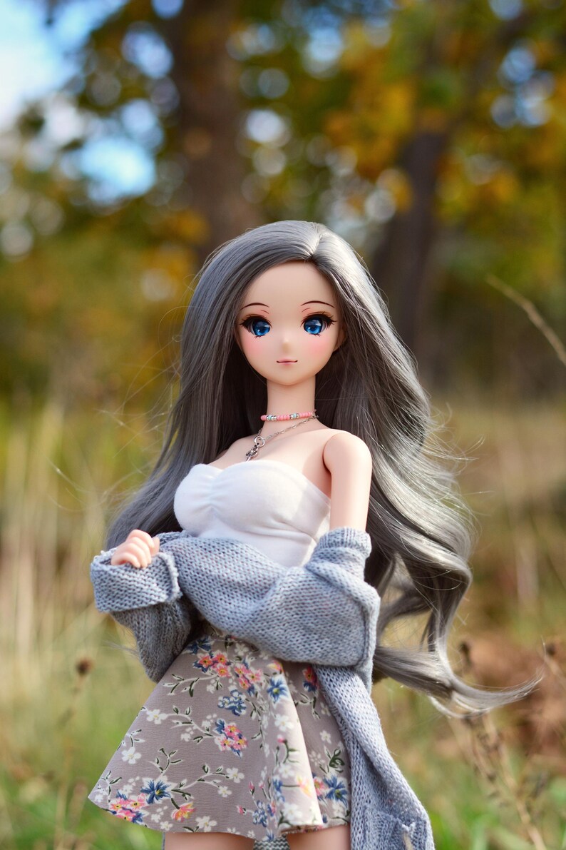 May include: A doll with long, gray hair wearing a white top, a floral skirt, and a light blue cardigan. The doll has blue eyes and is standing in a field with a blurred background of trees.