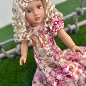 May include: A doll with blonde curly hair wearing a pink and brown floral dress. The doll has green eyes and is looking at the camera.